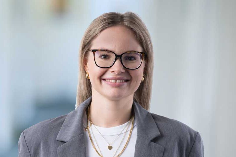 Junge Frau mit blonden, glatten Haaren und Brille lächelt in die Kamera. Sie trägt einen grauen Blazer, weißen Pullover und goldene Ketten. Der Hintergrund ist unscharf.
