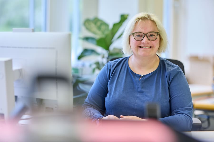 Lächelnde Frau mit blonden Haaren und Brille sitzt freundlich an ihrem Büroarbeitsplatz vor einem Computerbildschirm